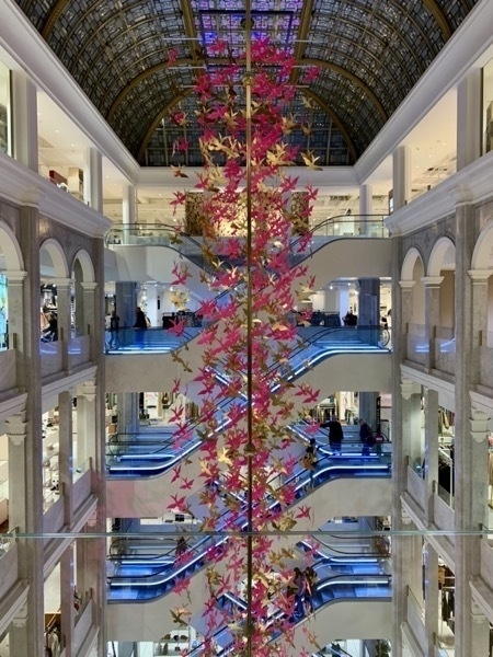 Interior of Illum department store in Copenhagen, with criss-crossing escalators in the background and an enormous column attached with red and gold birds suspended from the ornate stained-glass roof.