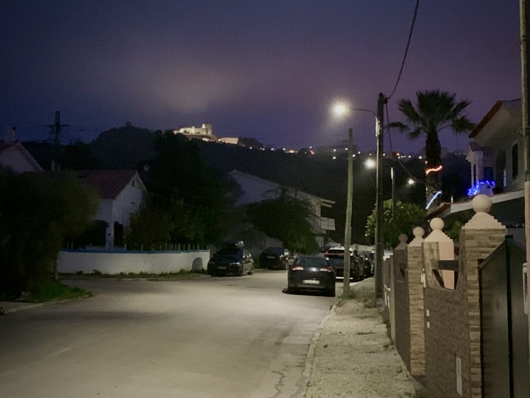 View of a residential street with the medieval castle of Palmela on a hill visible in the background, with lights illuminating the low clouds in the sky.