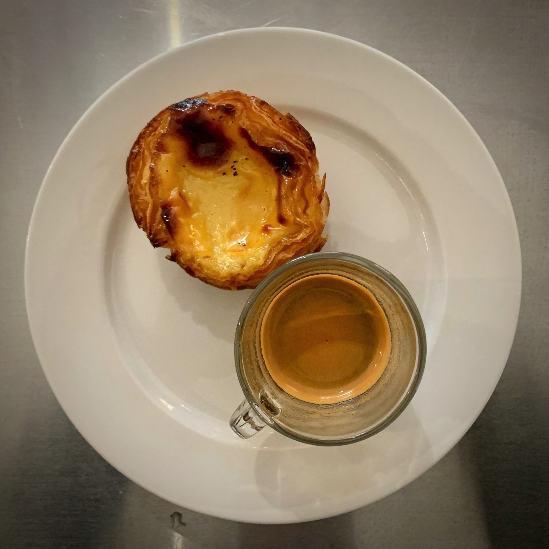 Espresso coffee in a glass cup and a Portuguese-style custard tart, set on a white plate